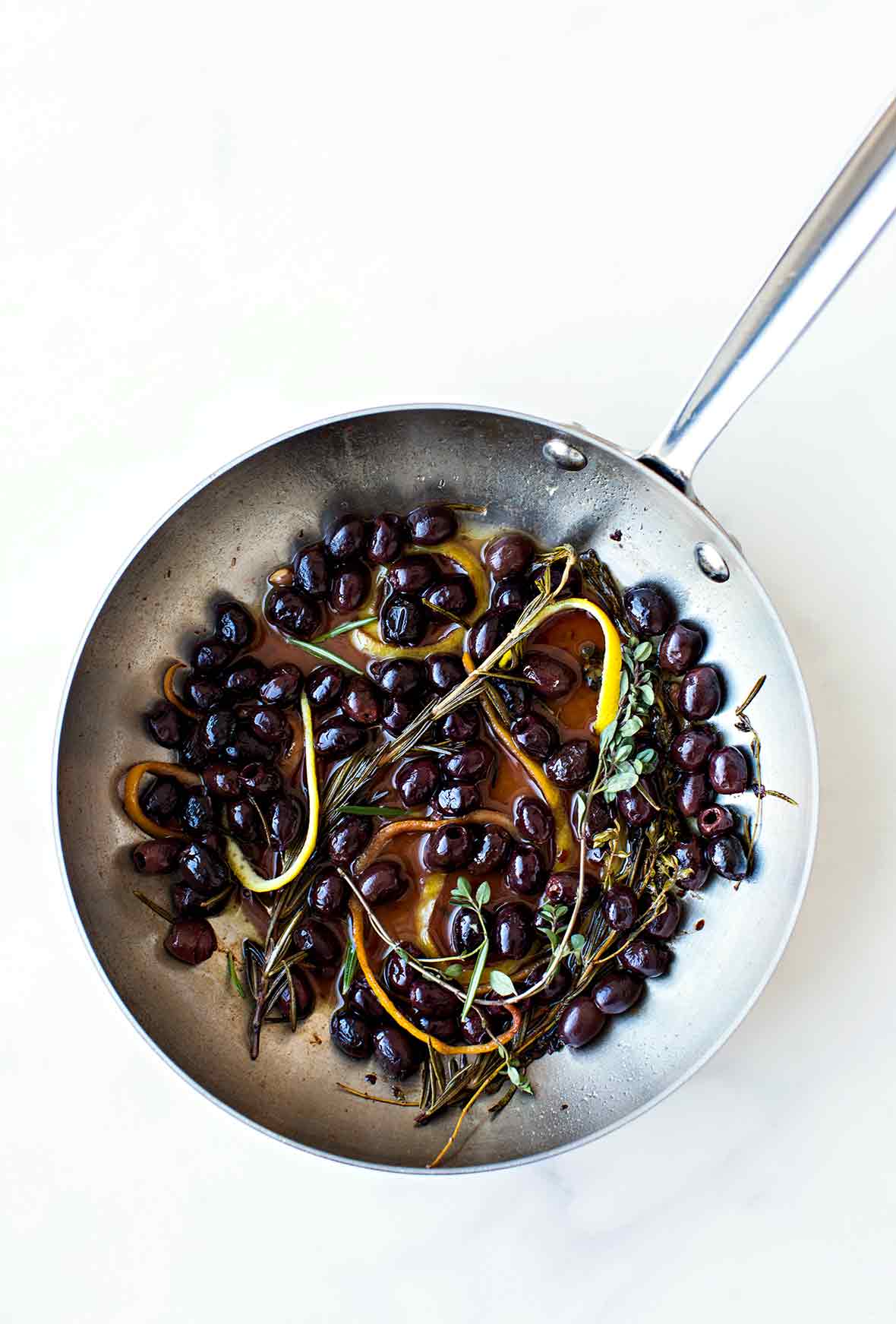 A skillet filled with black olives in an infusion of herbs and lemon and orange peel