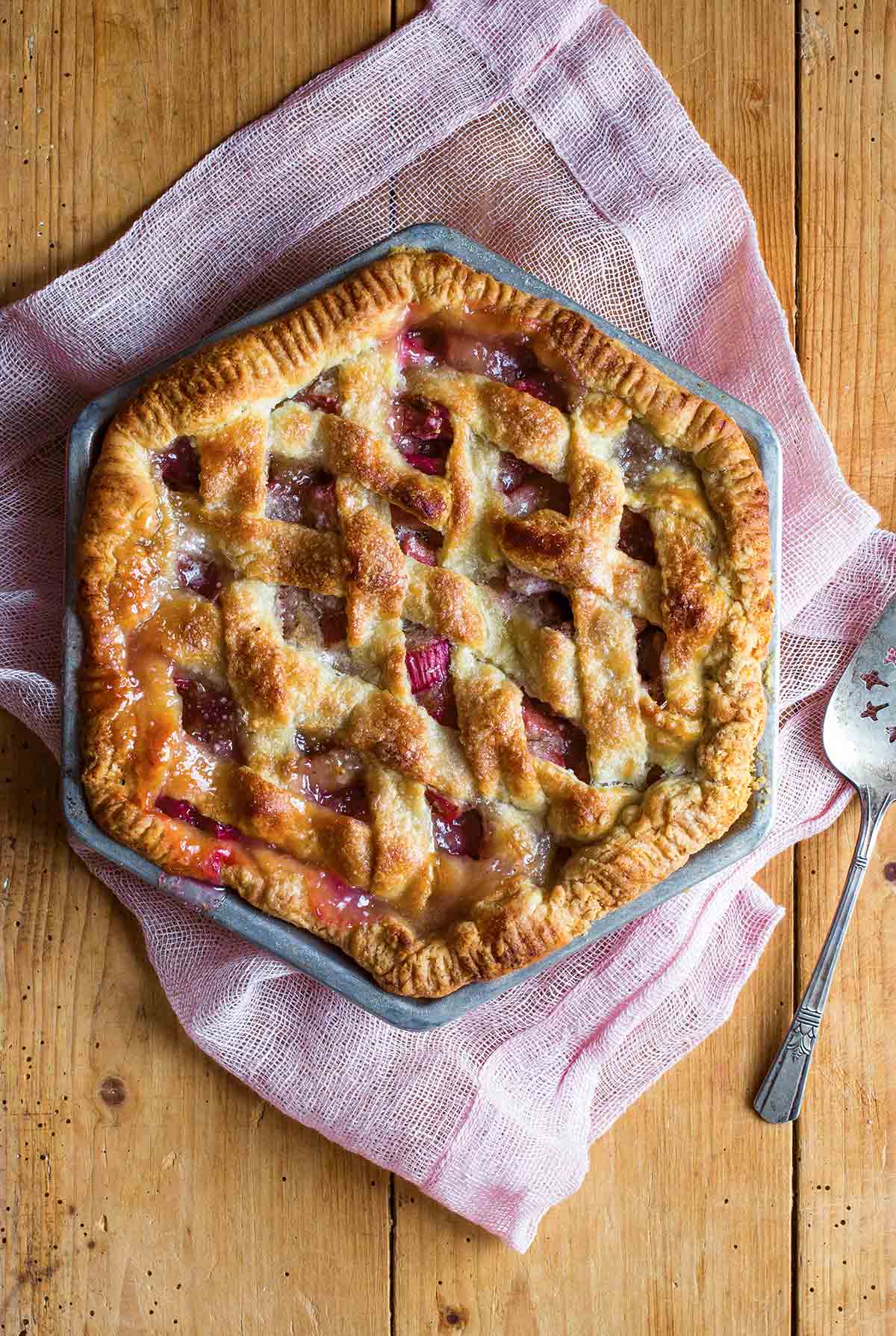 A hexagonal rhubarb pie with a lattice top crust on a pink linen cloth with a pie server resting beside the pie.