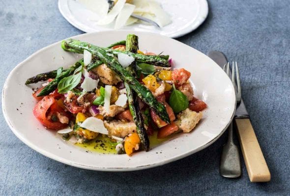 A white plate topped with a spring panzanella made with tomatoes, bread, asparagus, and Parmesan. A small plate of additional shaved Parmesan sits beside it.