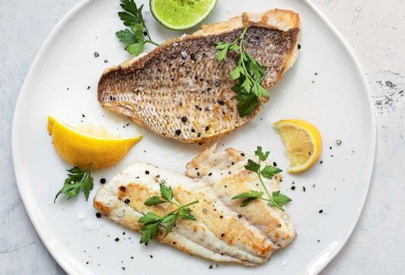 Two pieces of pan-seared fish fillet on a white plate with lemon wedges, a lime half, and parsley, to garnish.