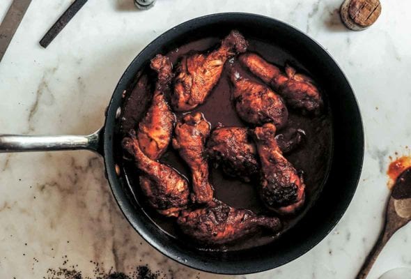 A skillet filled with chicken drumsticks braised in wine with a wooden spoon and wine cork lying beside the skillet.