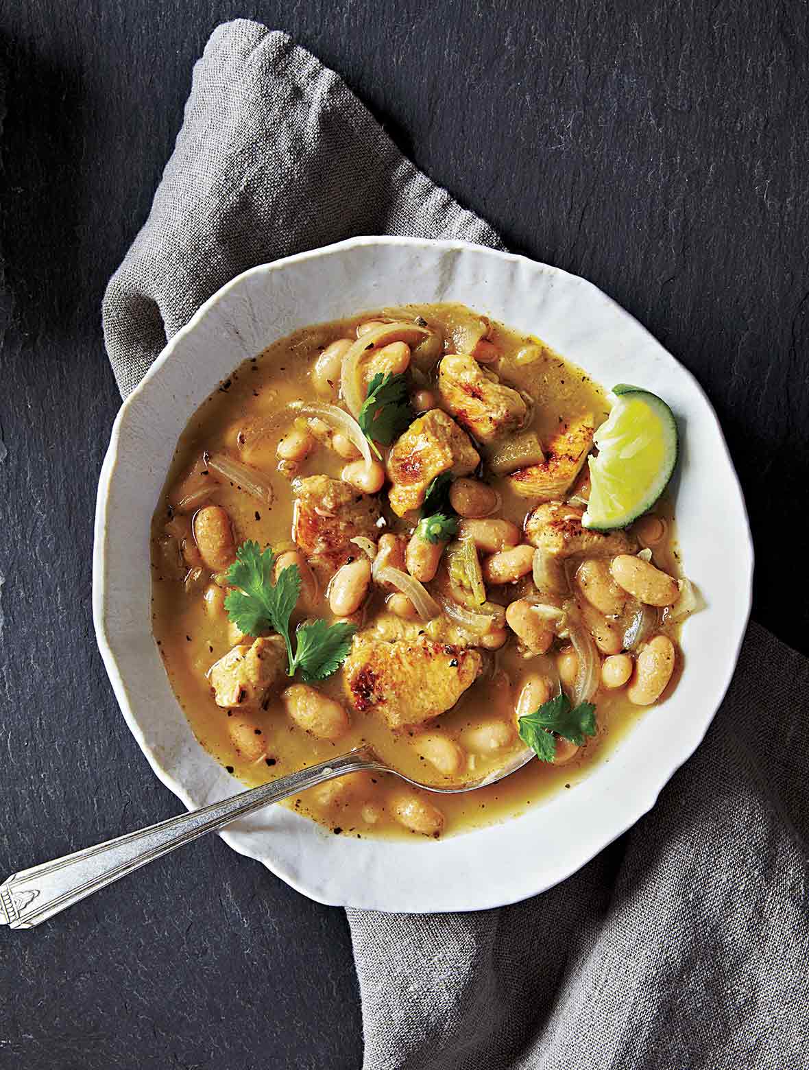 A white bowl on a grey linen napkin filled with white beans, chicken, onions, and topped with cilantro and lime