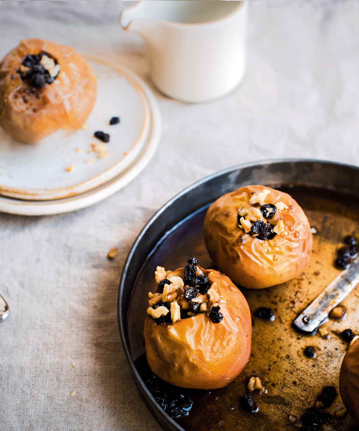 A metal baking dish with two baked apples in it, each apple topped with almonds and currants