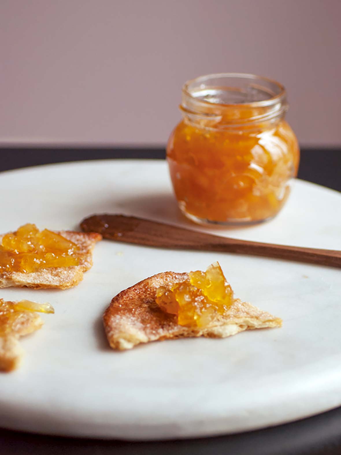 A serving platter with a piece of toast spread with marmalade. A jar of marmalade and knife rest in the background