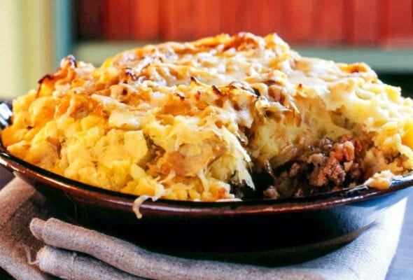 A porcelain bowl filled with shepherd's pie and topped with potatoes, onions, and cheddar
