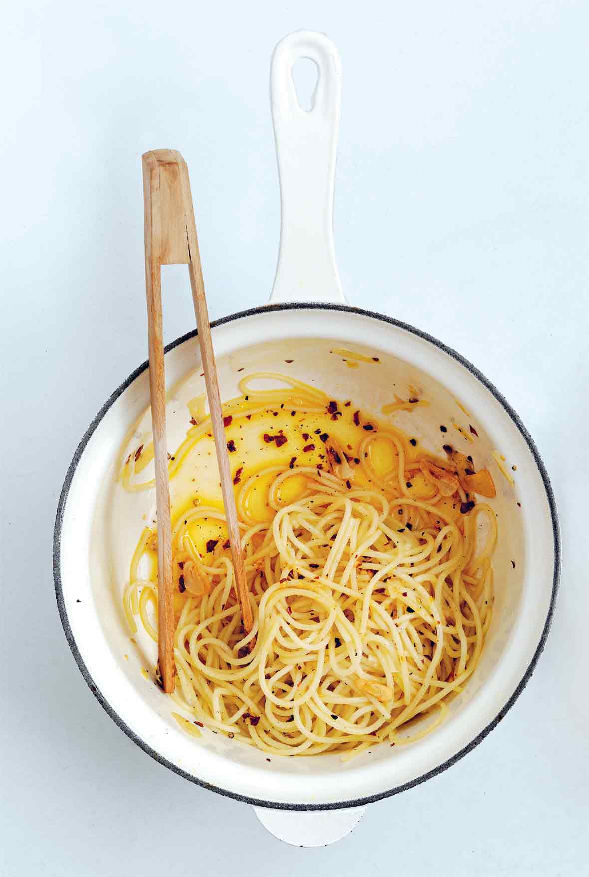 A white enamel pot with a blue rim with spaghetti, chili flakes, and oil in the bottom. A set of wooden tongs sits inside.