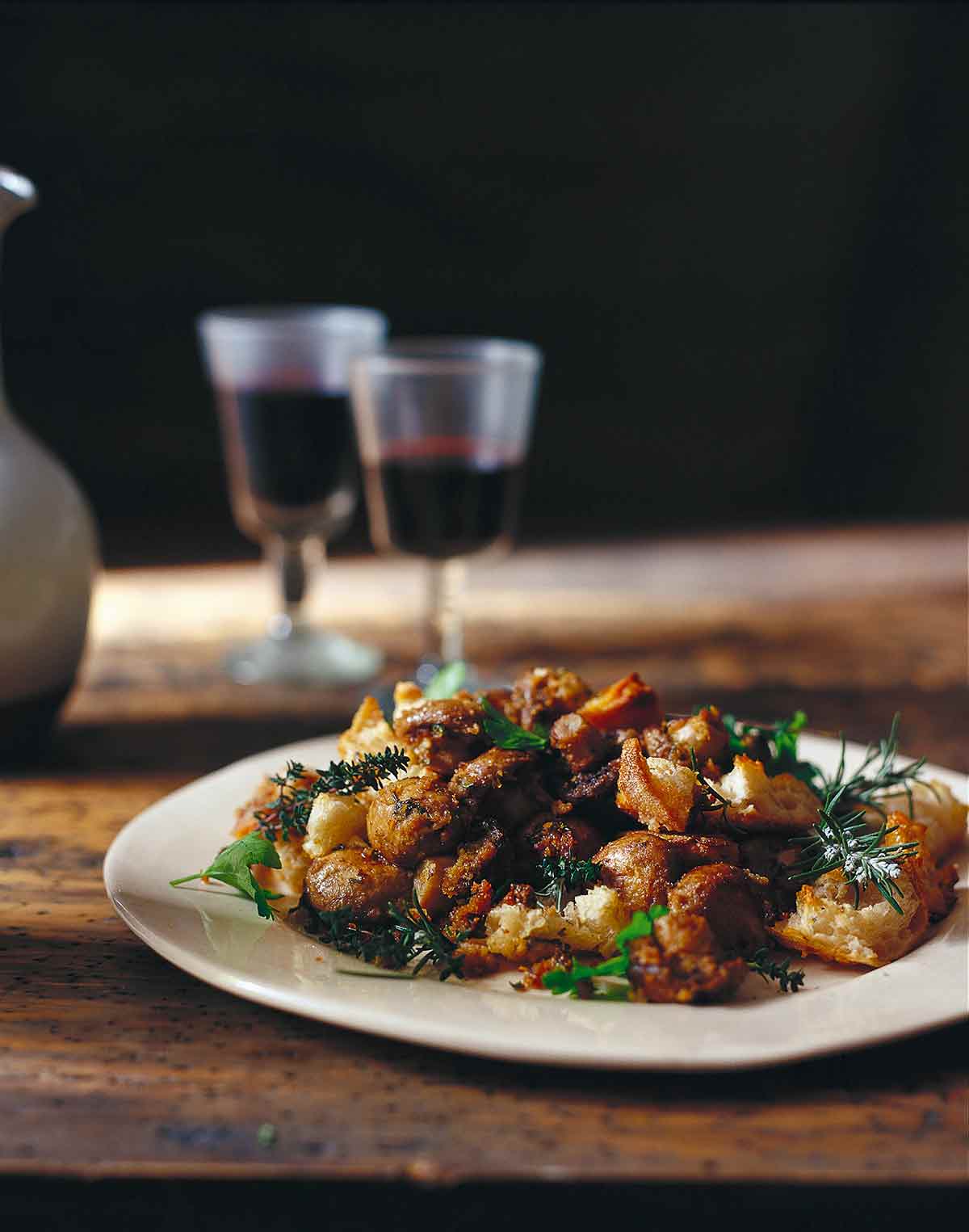 A white plate filled with baked mushrooms, broken bread, and two glasses of red wine in the background.