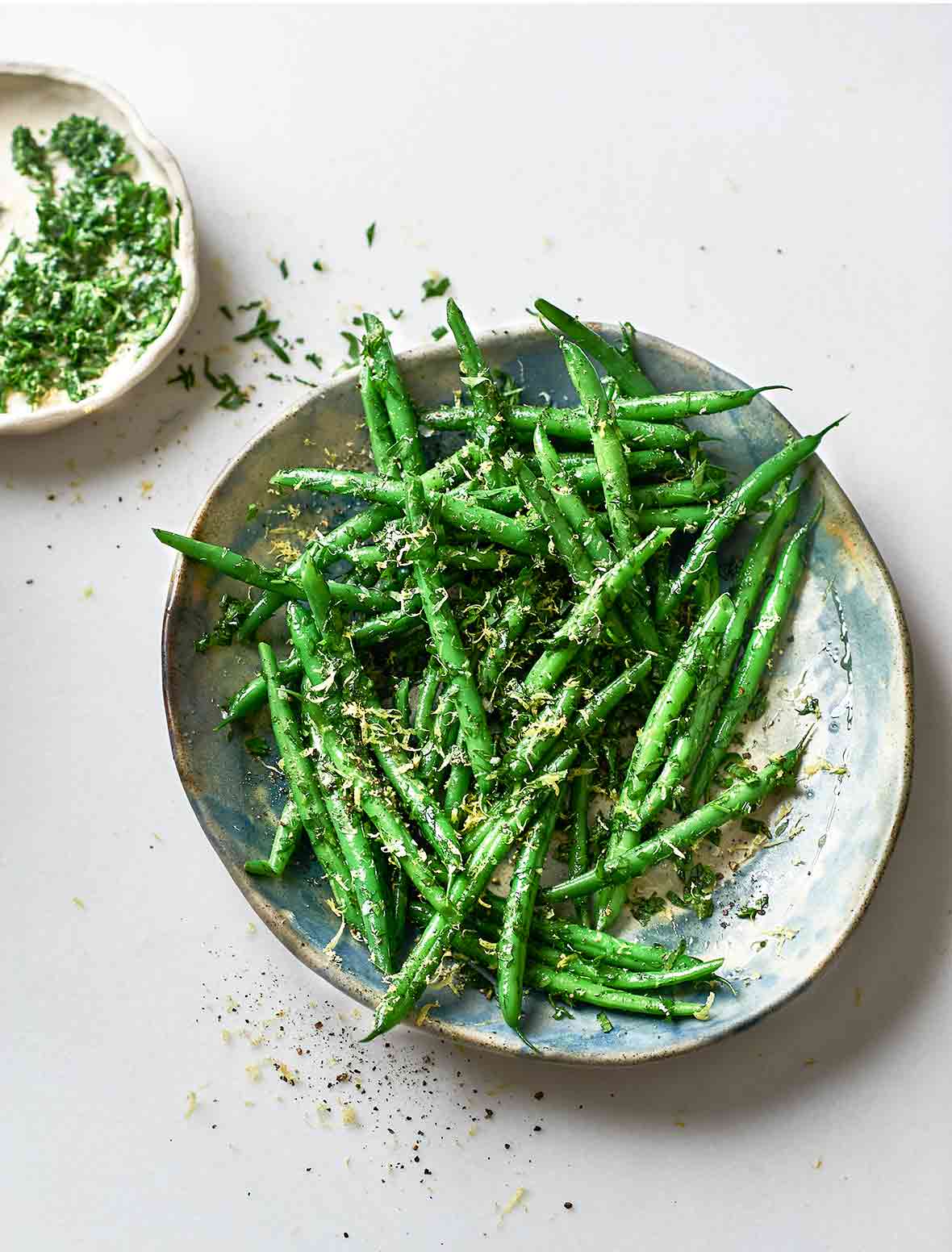 A serving platter piled with green beans and sprinkled with gremolata. A small dish of gremolata sits beside the serving plate.
