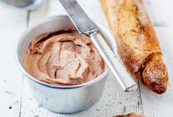 A metal bowl filled with brown chicken liver pate; a knife balances on the edge; nearby is a baguette