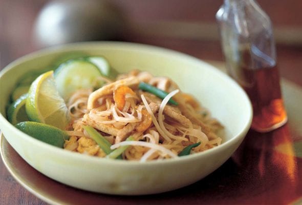 Pad Thai noodles, crunchy noodles. cucumber, bean sprouts, and lime wedges in a bowl, a bottle of chile-vinegar