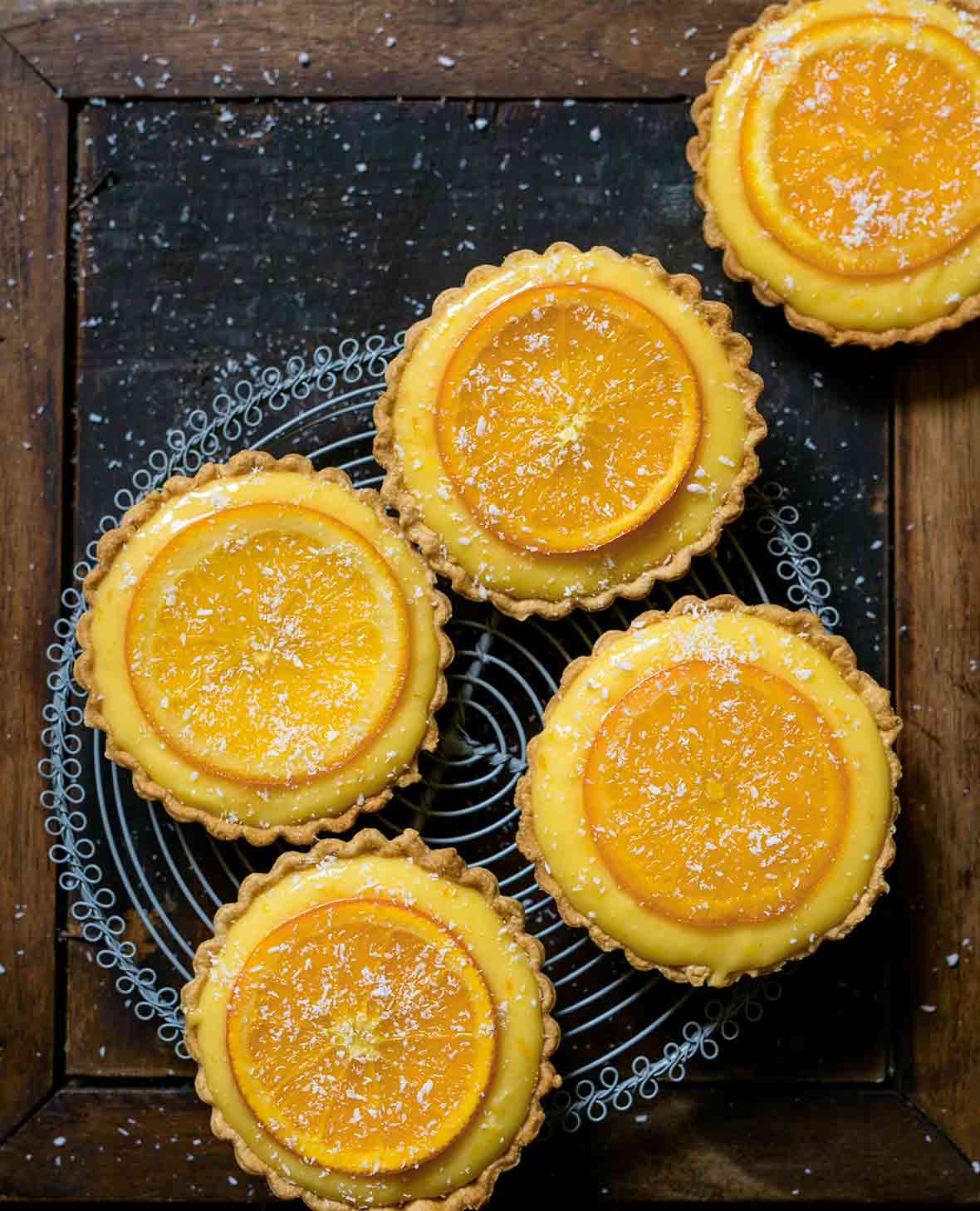 Five small tarts filled with orange curd and topped with candied orange slices on a cooling rack