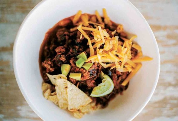 White bowl of beef chili with grated cheese, tortilla chips, and lime wedges on wood