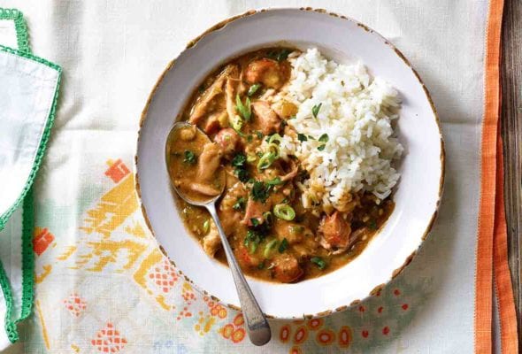 A chipped white bowl filled with Cajun chicken and sausage gumbo and a scoop of rice with a spoon resting in it.