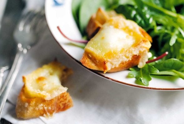 A bowl filled with baby greens on a white linen napkin. Part of a goat cheese crostini is on the edge of the bowl and the other part on the napkin