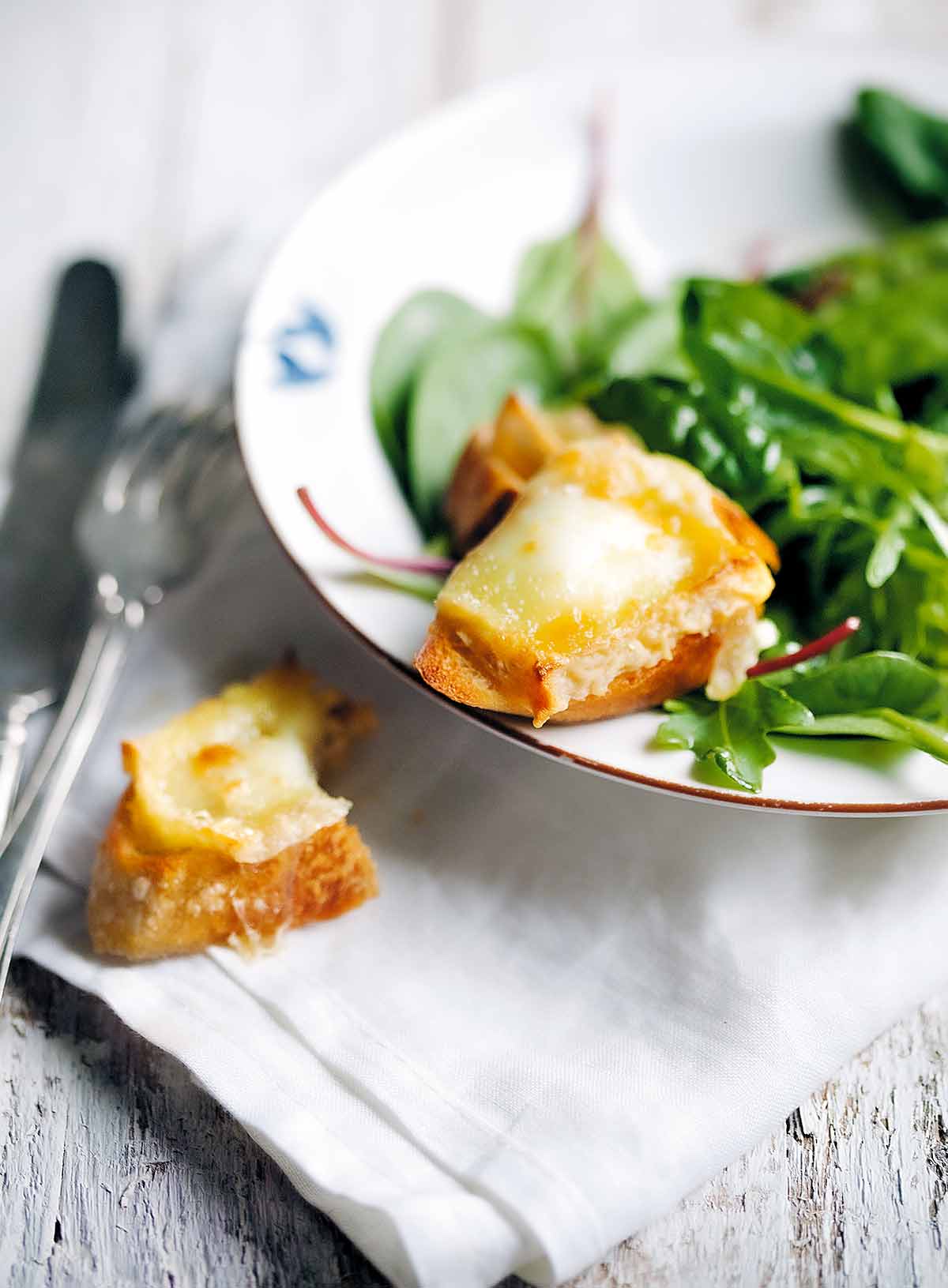 A bowl filled with baby greens on a white linen napkin. Part of a goat cheese crostini is on the edge of the bowl and the other part on the napkin