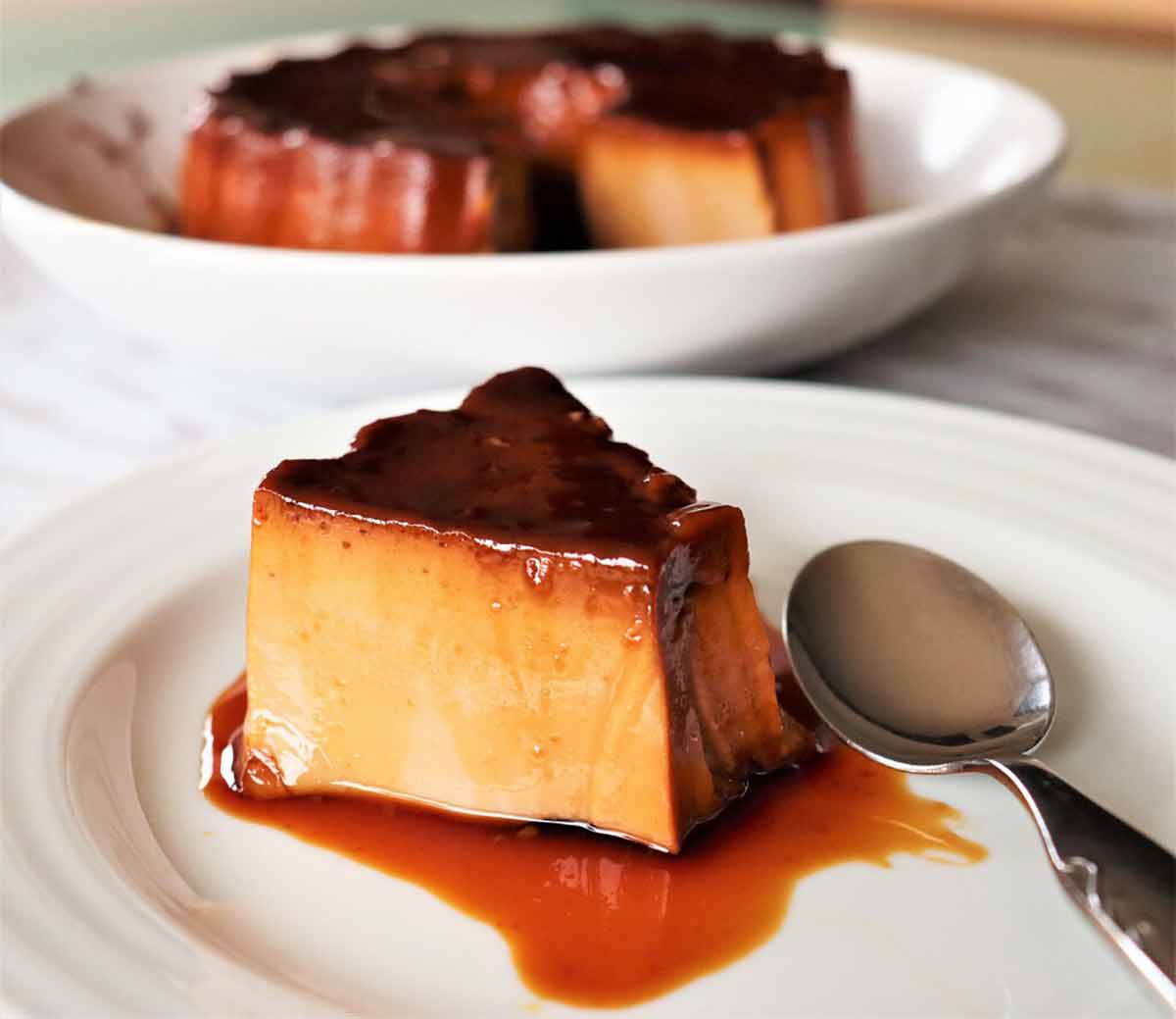 A piece of Portuguese tea flan on a white plate, drizzled with caramel sauce with the remaining flan in a bowl in the background.