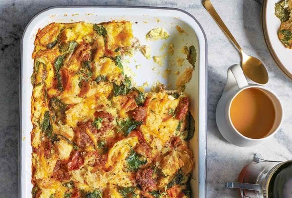Baking dish of sausage egg casserole of bread, eggs, sausage, tomato, kale, and Cheddar cheese, a plate and tea nearby