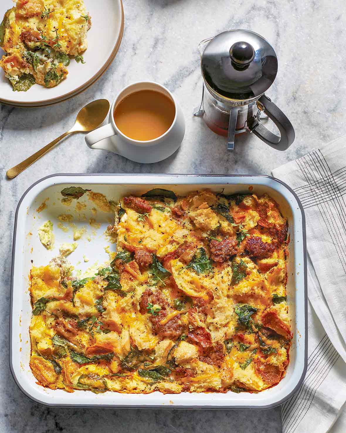 Baking dish of sausage egg casserole of bread, eggs, sausage, tomato, kale, and Cheddar cheese, a plate and tea nearby