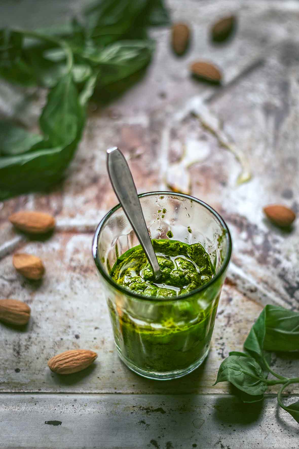 Glass of spinach almond pesto with a spoon inside on a metal sheet strewn with almonds