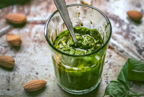 Glass of spinach almond pesto with a spoon inside on a metal sheet strewn with almonds