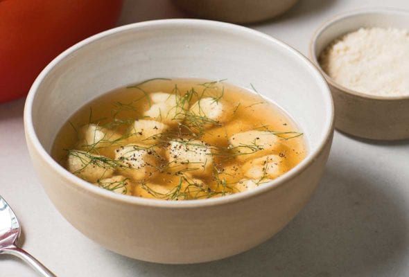 A bowl of chicken soup with dumplings, topped with fennel fronds, along with red pot and bowls