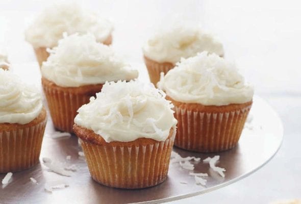 Cake stand with 7 frosted coconut cupcakes, four unfrosted cupcakes on the table, a bowl of frosting