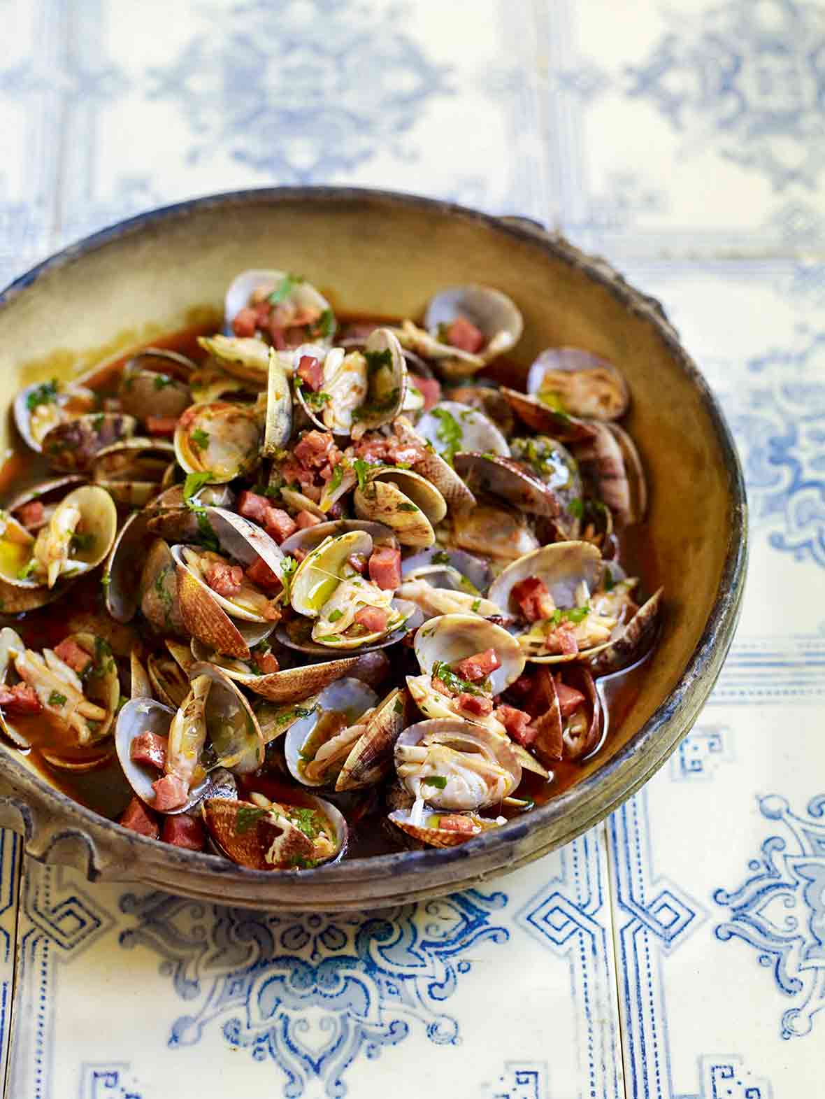 A bowl of clams with chouriço, garlic, and cilantro with a savory broth on top of blue and white tiles