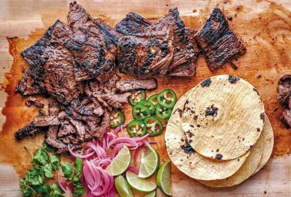 Cutting board with grilled skirt steak tacos, sliced jalepenos, tortillas, lime wedges, pickled onions, cilantro