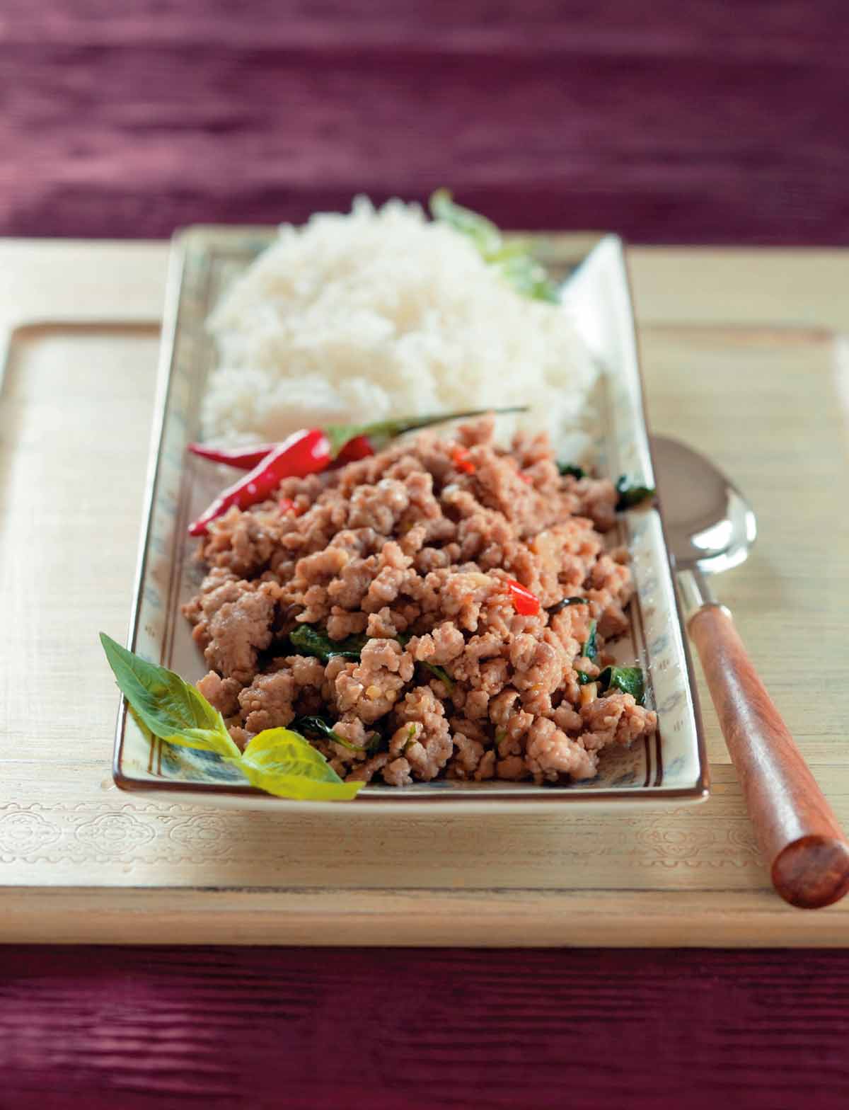 A rectangular serving dish filled with Thai basil pork, red chiles, and white rice with a serving spoon beside the dish.