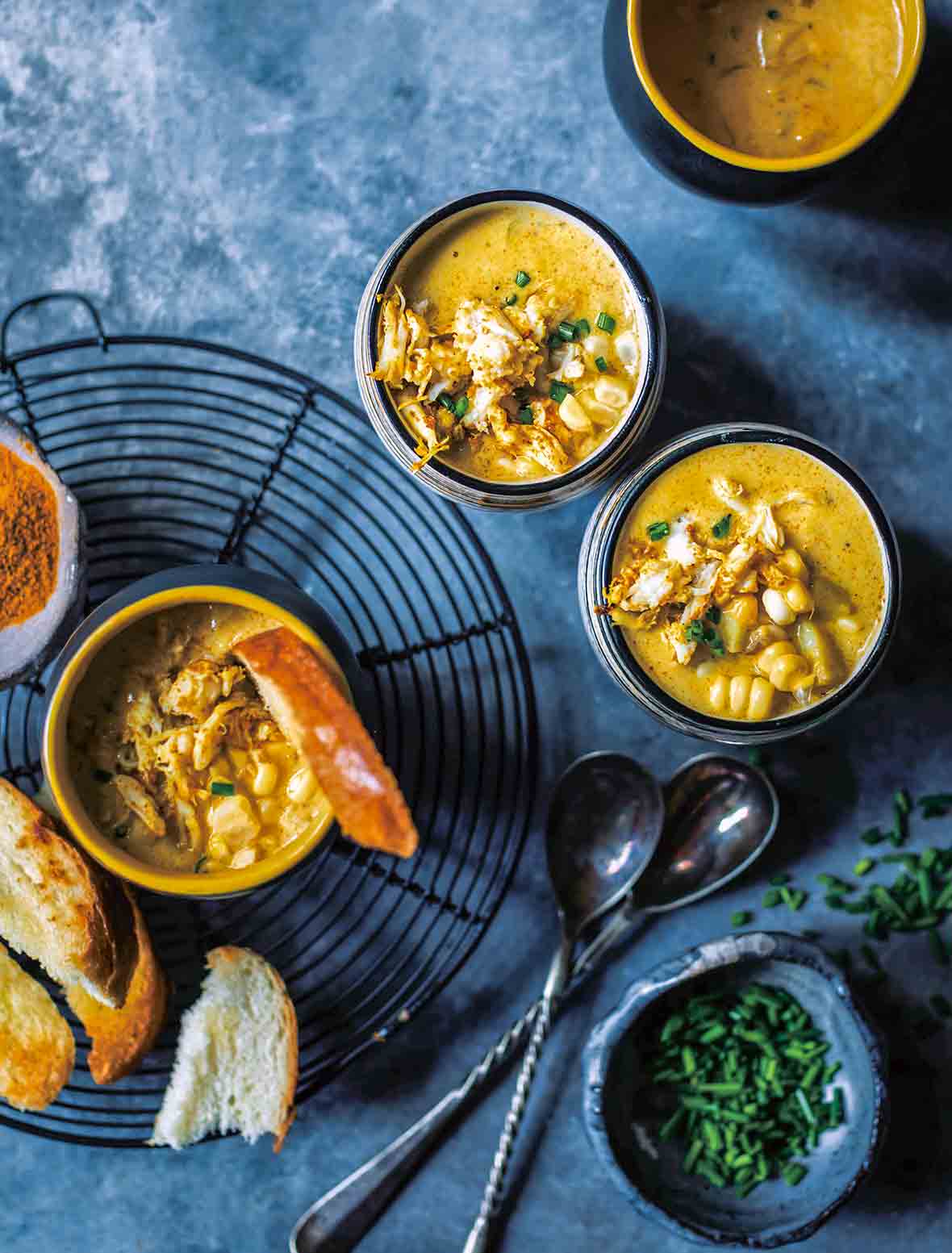Four bowls of curried crab corn chowder, filled with crab, corn, and a curried broth, on bread and spoons nearby