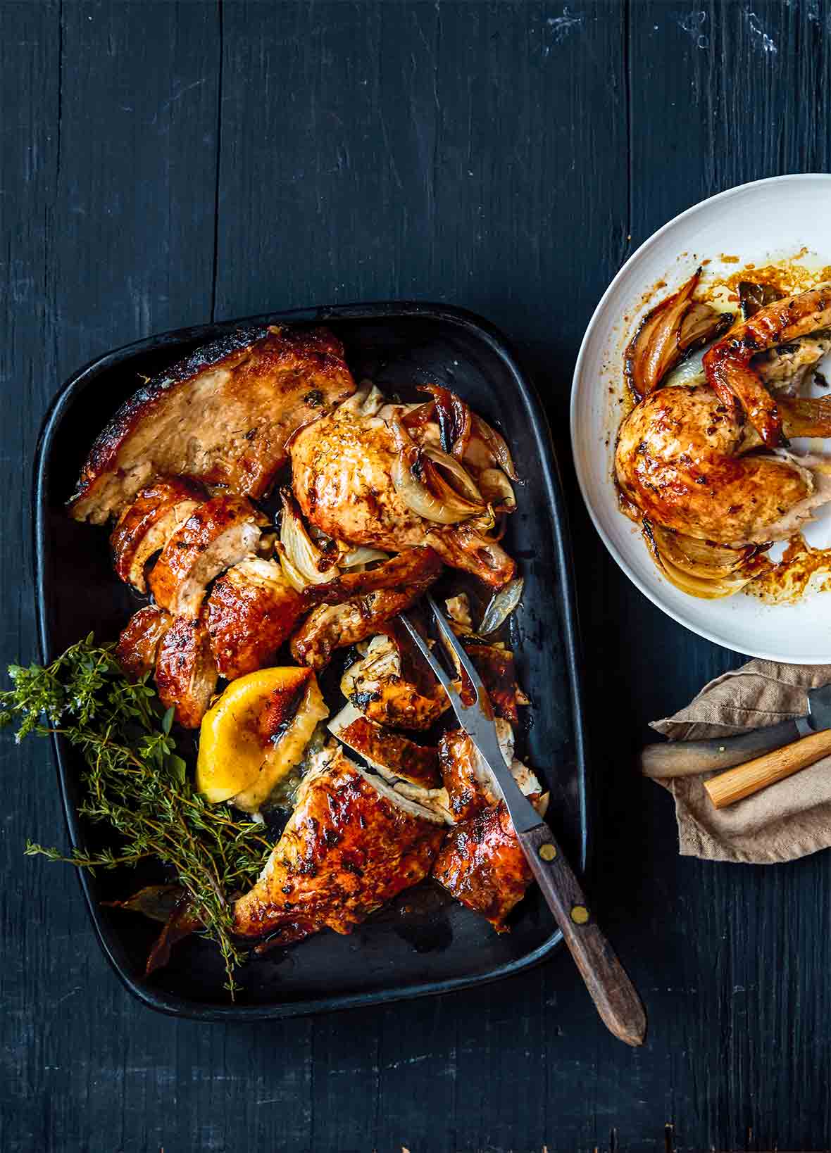 A black platter with golden brown herb butter chicken, roasted onion, a lemon wedge, and sprigs of thyme and oregano by Dorie Greenspan