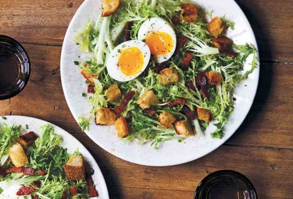 Two white plates of frisée with bacon and egg, croutons, and a dressing, glasses and fork and knife nearby
