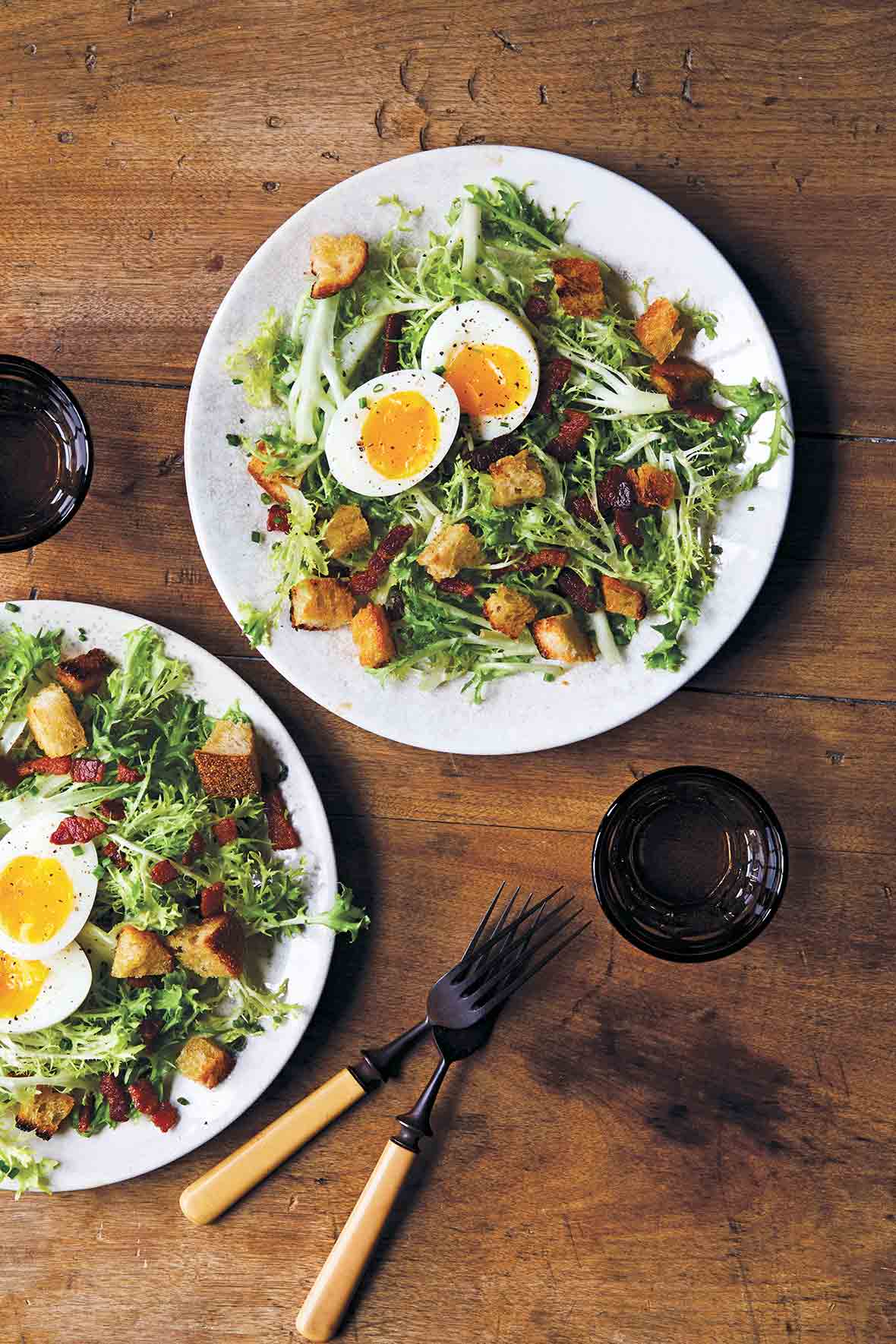 Two white plates of frisée with bacon and egg, croutons, and a dressing, glasses and fork and knife nearby