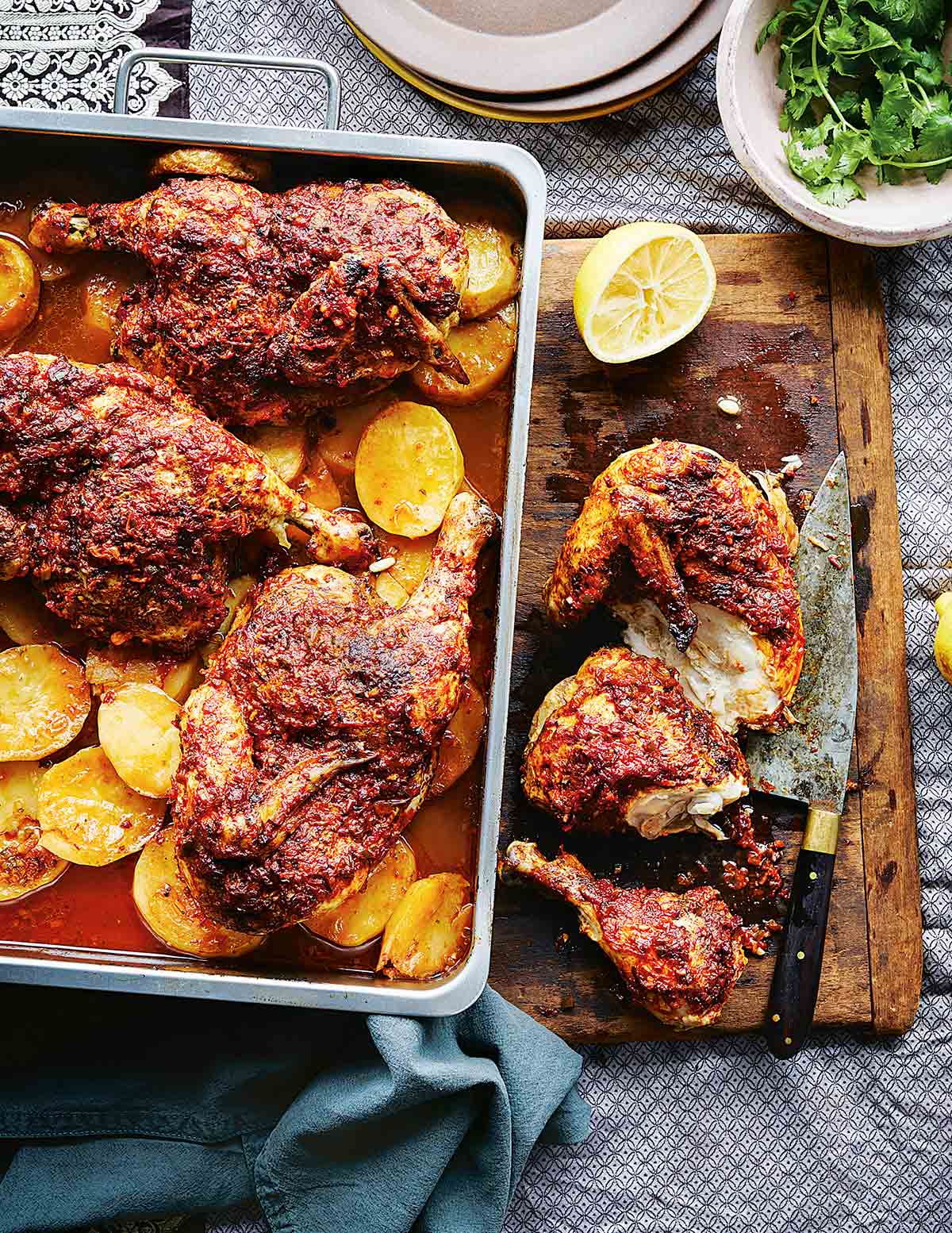 A pan of harissa roasted chicken sitting on top of sliced roasted potatoes, nearby a cutting board and knife