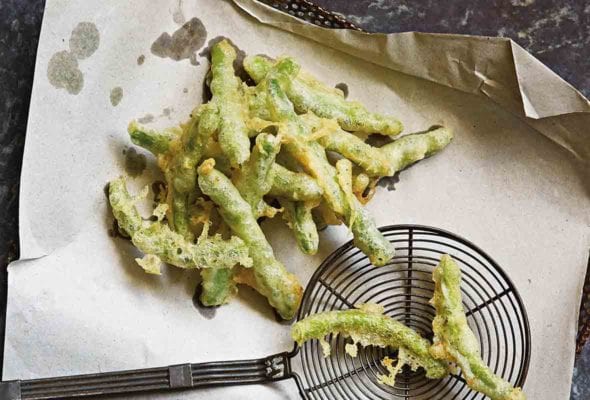 A pile of Portuguese peixinhos da horta (tempura green beans) draining on brown paper in a strainer, a spider spatula nearby