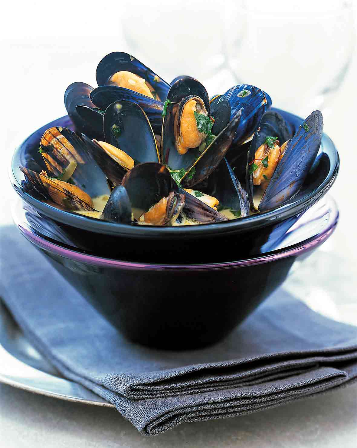 A bowl of pressure cooker mussels with saffron sprinkled with chopped parsley and in a yellow broth set in another bowl on top of a napkin