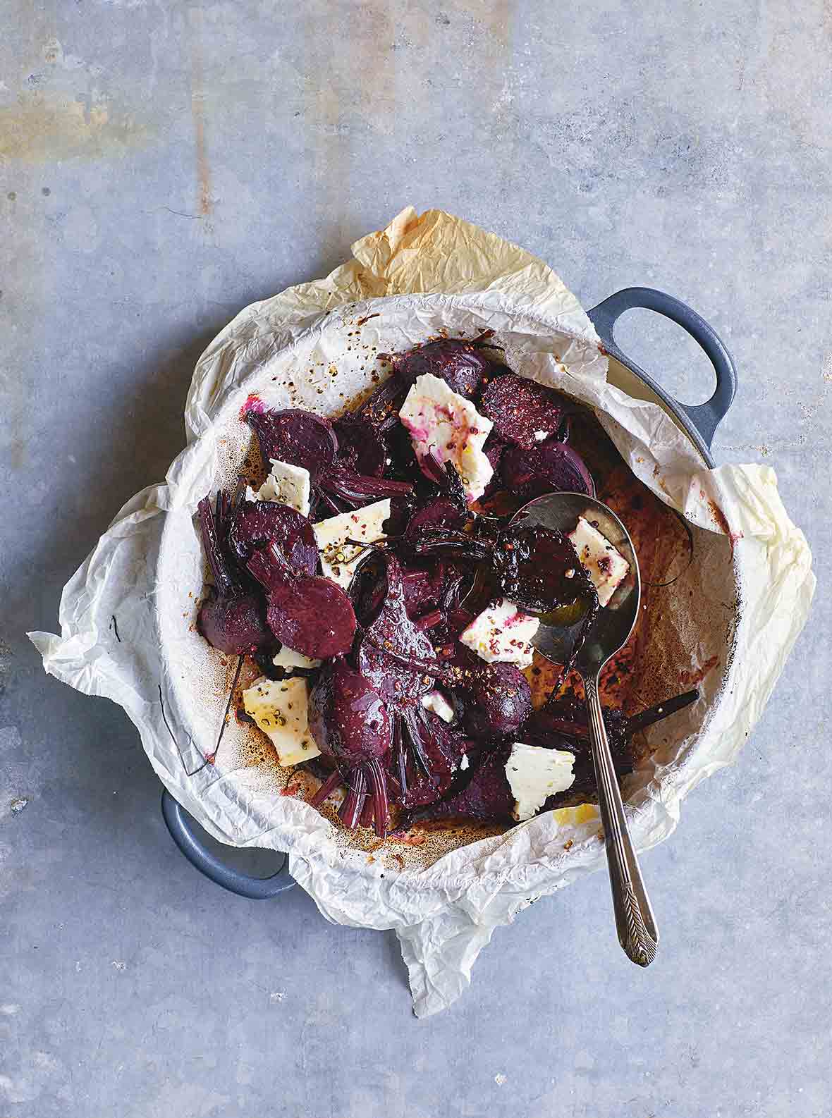 A pan lined with parchment paper containing sliced roasted beets, chunks of feta cheese, a dressing, and a spoon