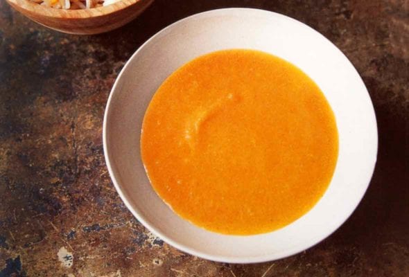 A white bowl filled with carrot and lentil soup and a wooden bowl of cooked rice beside it.