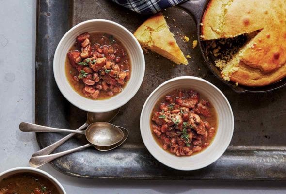 Three white bowls of pork and pinto beans chili on a metal tray with cornbread in a skillet