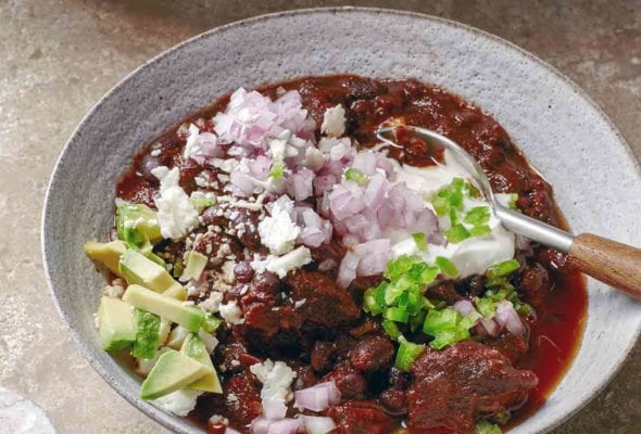 A white bowl filled with beef and black bean chili, topped with red onion, avocado, queso fresco, and diced jalapeno