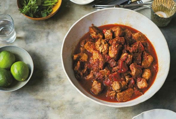 A bowl of pork tinga--pork stewed with tomatoes and chipotle peppers, along with bowls of lemon, sour cream, cilantro, cheese on a table