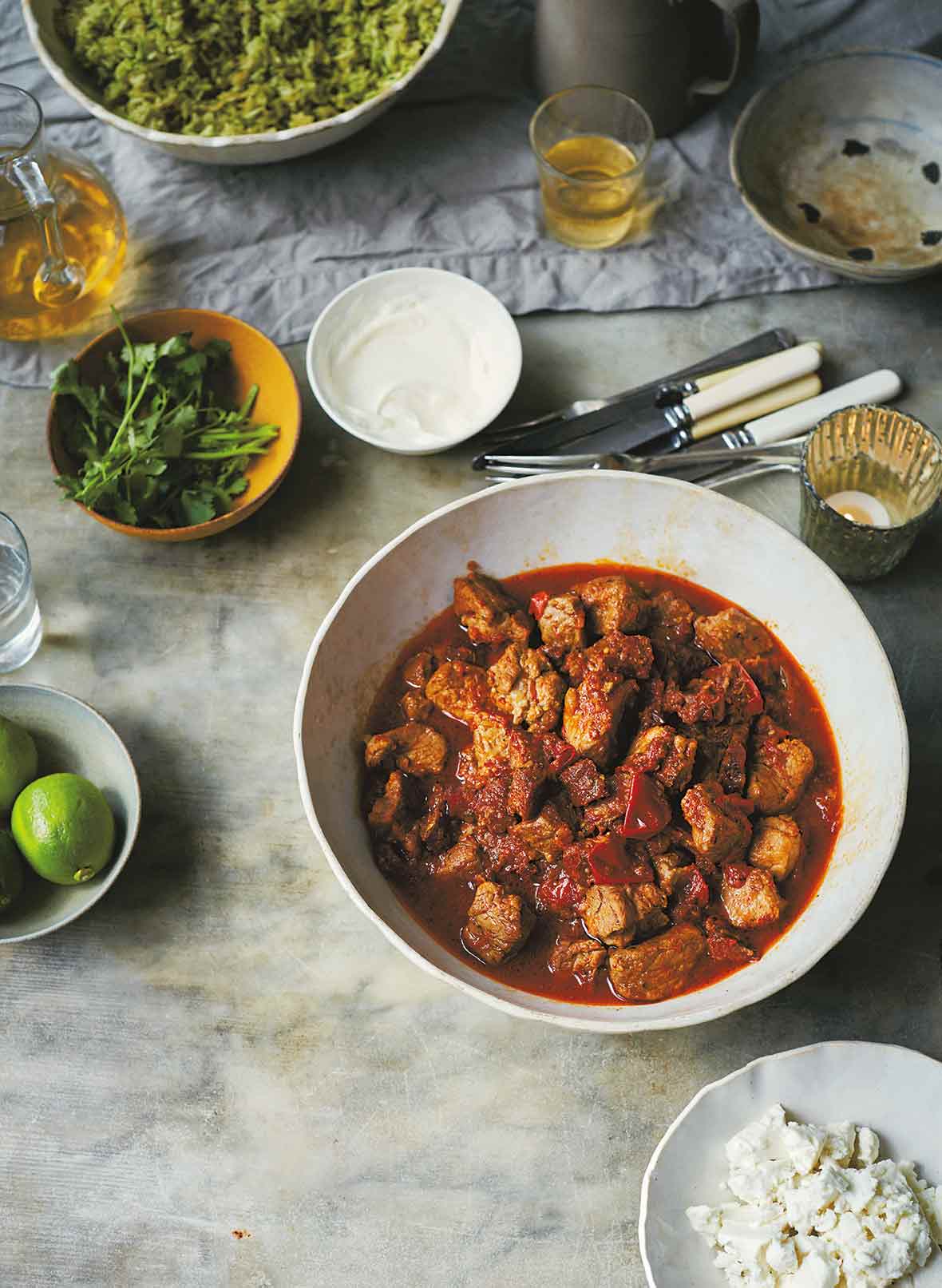 A bowl of pork tinga--pork stewed with tomatoes and chipotle peppers, along with bowls of lemon, sour cream, cilantro, cheese on a table