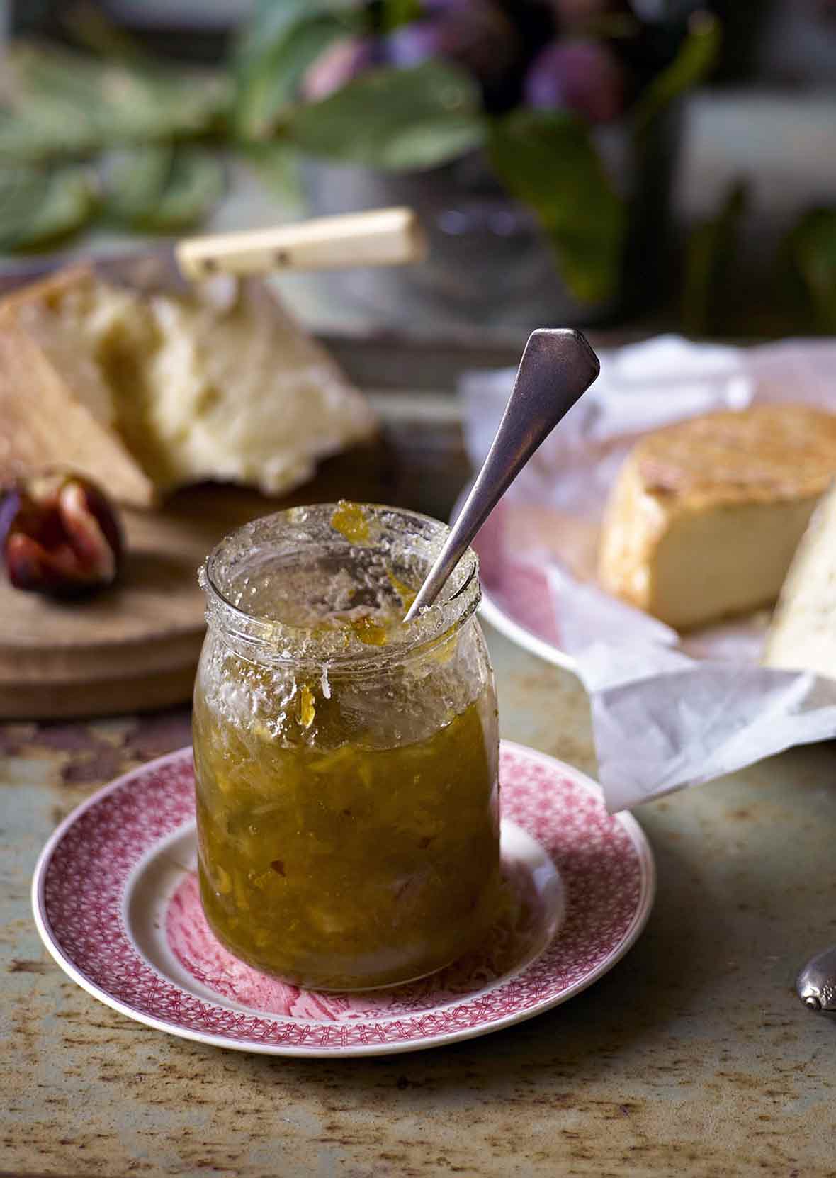 A jar of celery marmalade with a spoon in it and several types of cheese in the background