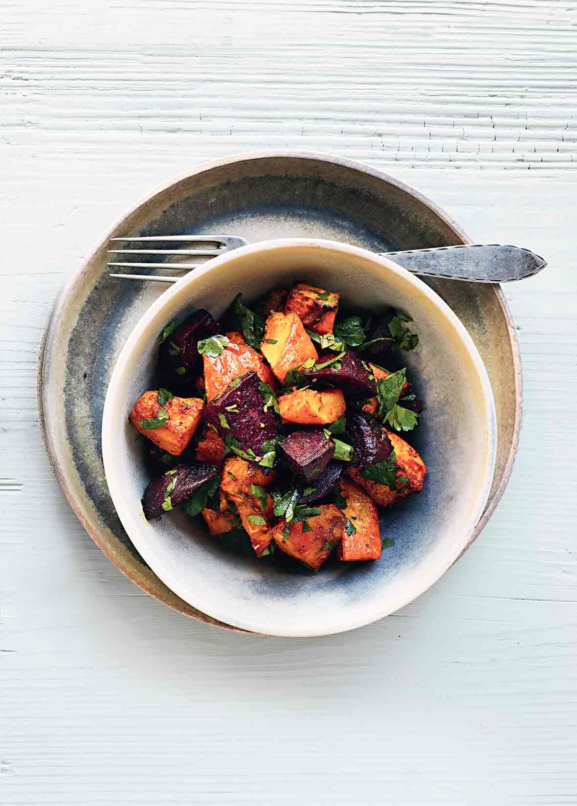 An earthenware bowl filled with curried butternut squash and beets, on a plate with a fork resting beside the bowl