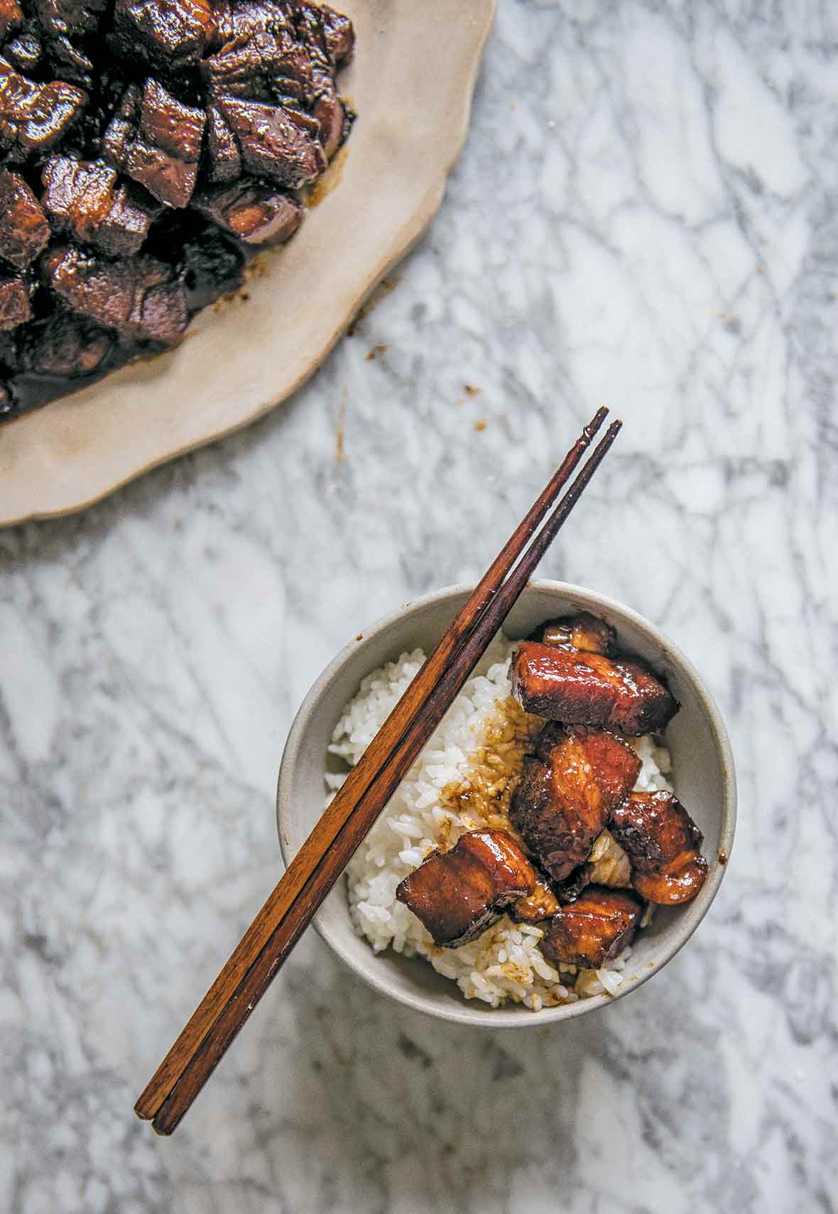 A bowl of rice topped with red cooked pork and a pair of chopsticks resting on the bowl. A plate of red cooked pork on the side