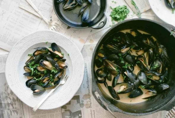 A pail and a white bowl filled with red curry mussels, as well as a bowl of discarded mussel shells