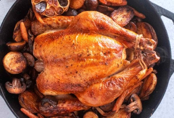 A best brined roast chicken in a cast iron Dutch oven surrounded by potatoes, mushrooms, carrots, and a halved head of garlic.