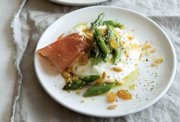 Two plates with burrata topped with asparagus, pine nuts, raisins, and prosciutto.