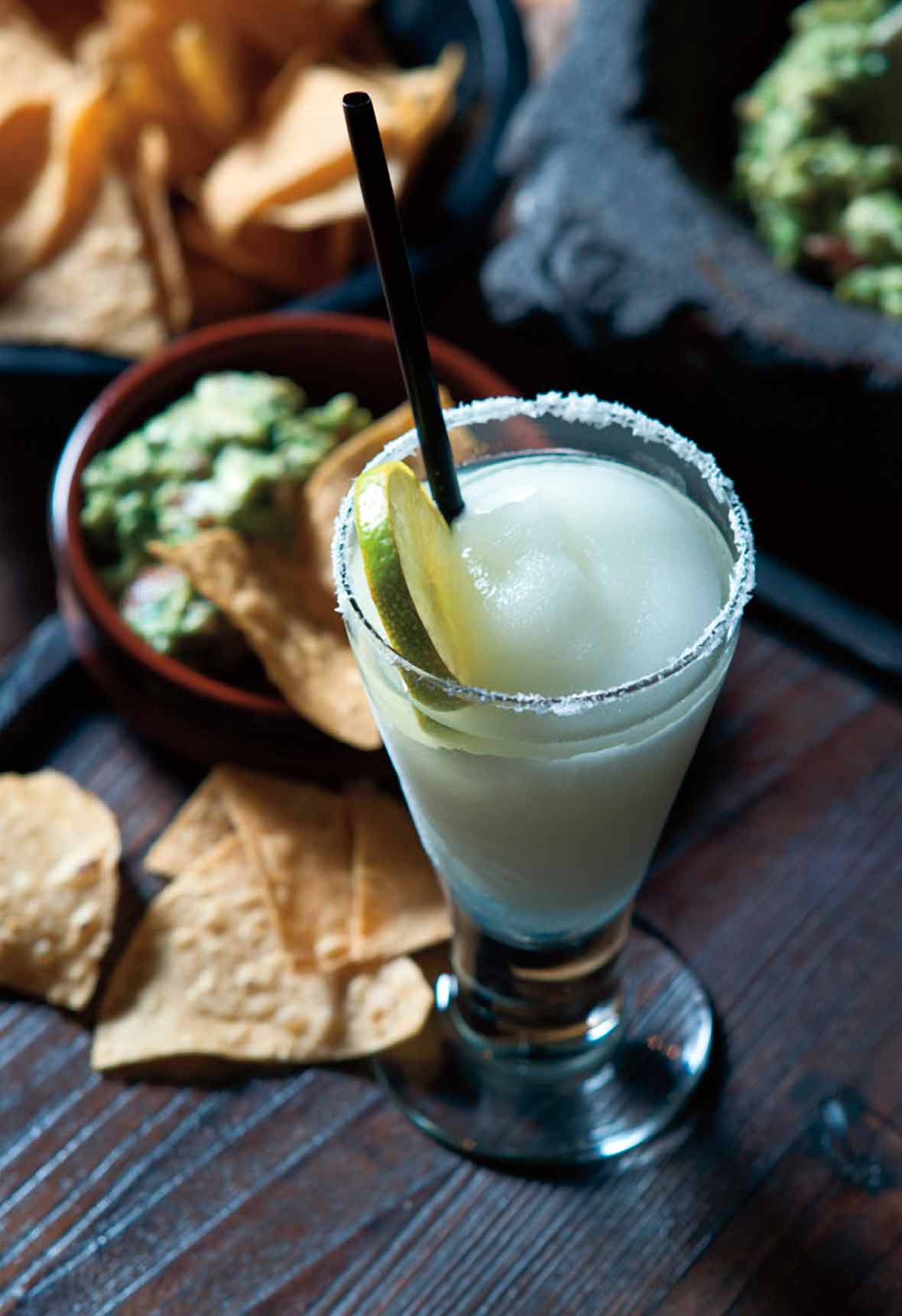 A tall glass of frozen margarita with a salt rim and lime wheel beside a bowl of chips and guacamole