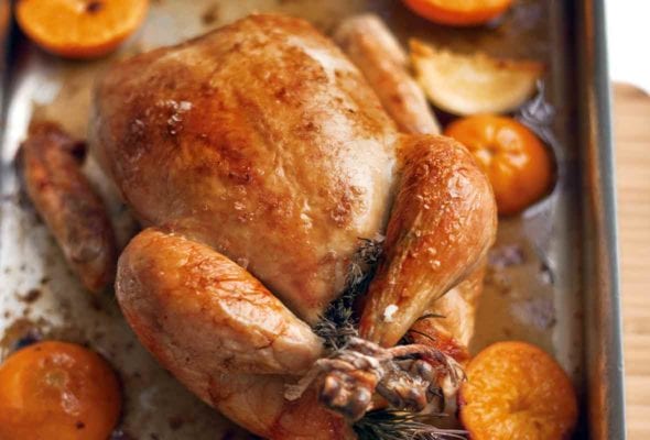 A whole roast chicken in a roasting pan with halved citrus fruits around it
